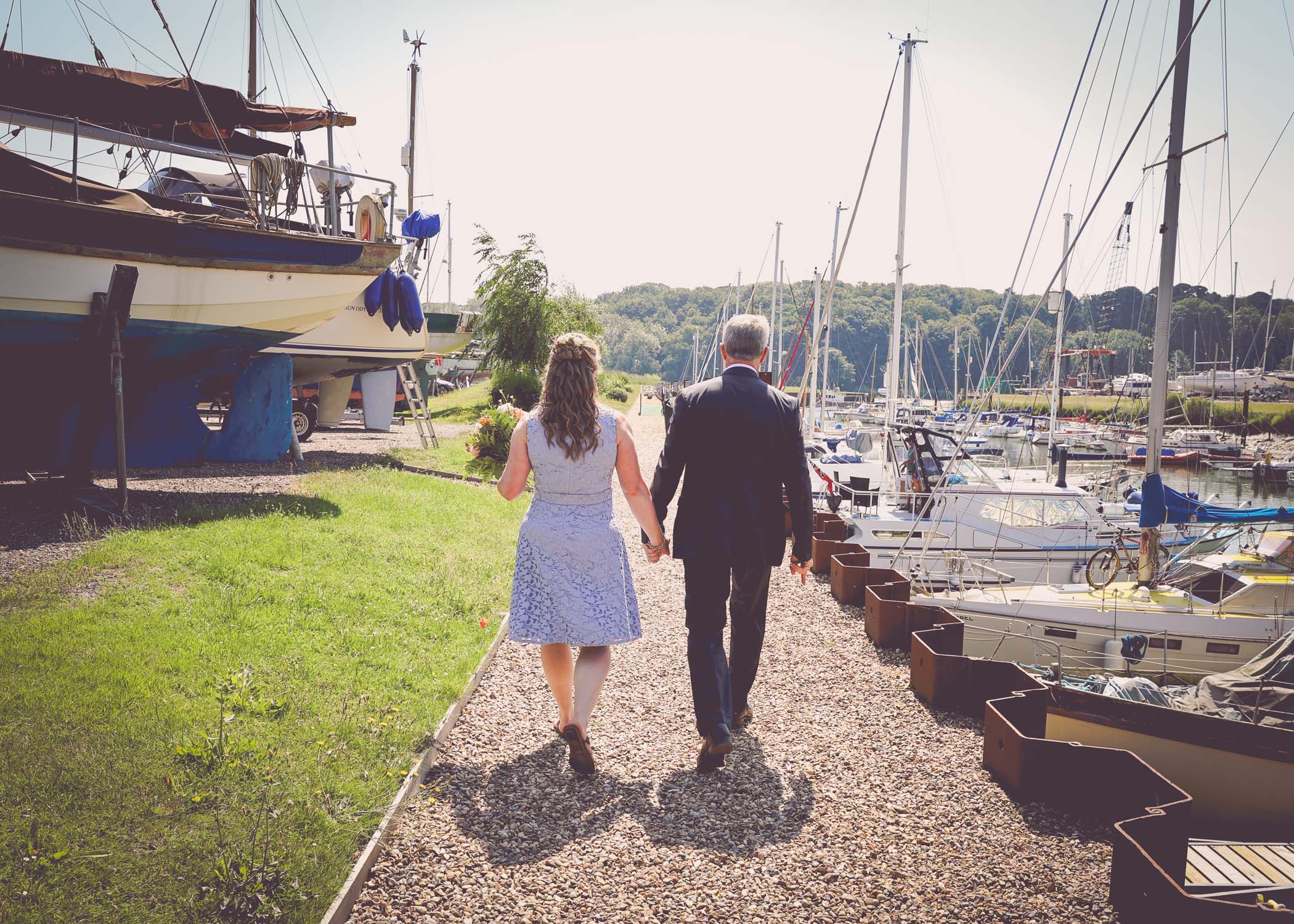 A bride and groom walking from behind through the yaughts in the marina on a wedding day in Woodbridge captured by Suffolk Wedding Photographer Hayley Denston Photography
